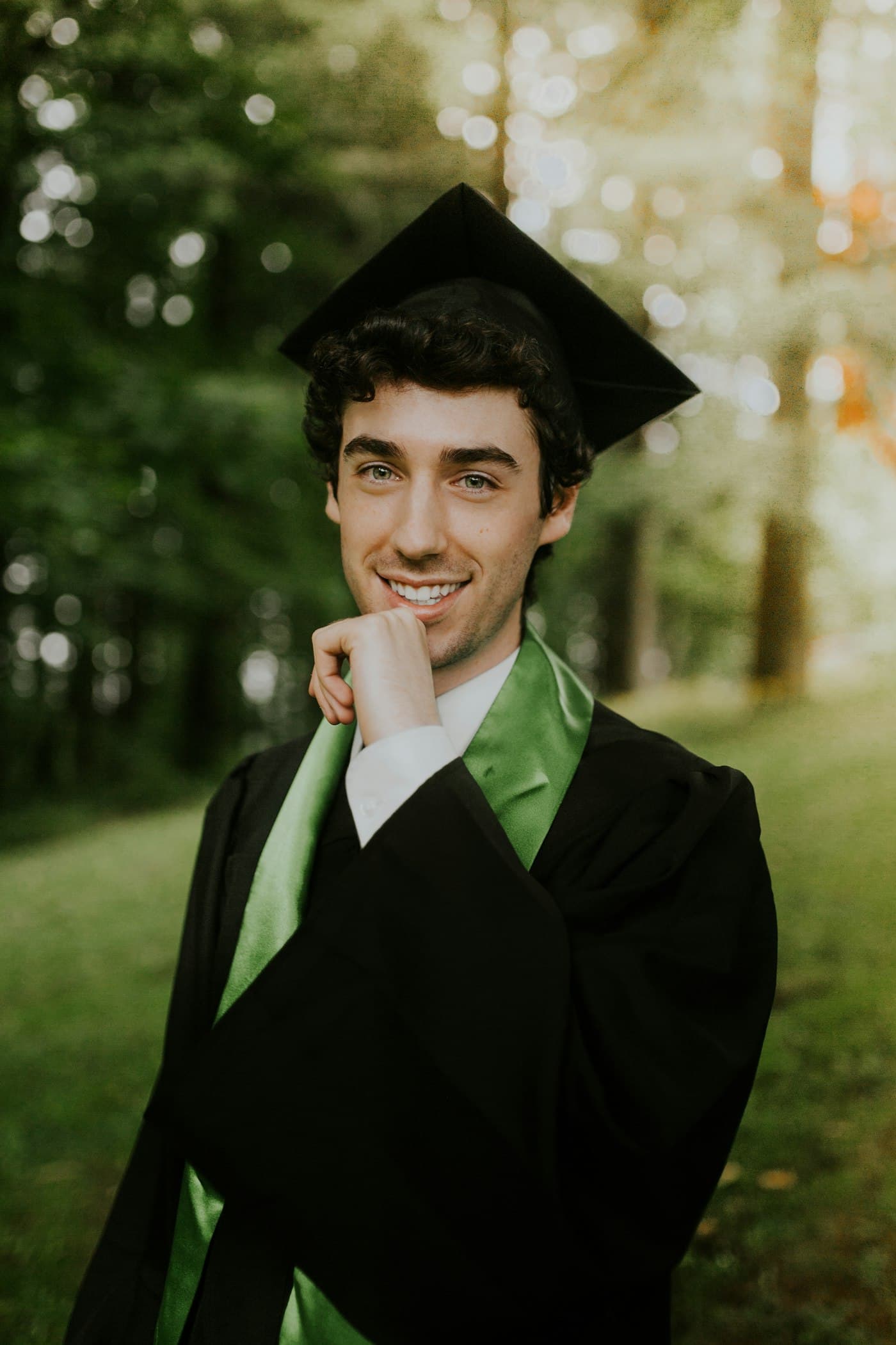 Wisudawan bertoga tersenyum dengan tangan di dagu di taman